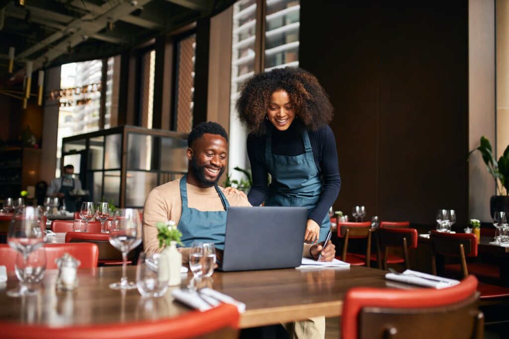 Dos propietarios de restaurante sonrientes revisan estrategias de marketing digital en una laptop, dentro de un comedor moderno y luminoso.